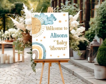 Könnte beinhalten: Willkommen zur Babyparty von Alisons! Ein weißes Schild mit goldenen und blauen Aquarell-Illustrationen einer Sonne, Wolken und eines Regenbogens. Das Schild steht auf einem Holzstaffelei.