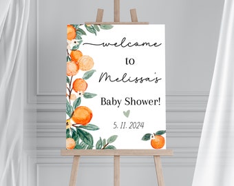May include: A white sign on a wooden easel with the text "welcome to Melissa's Baby Shower!" in black script. The sign is decorated with watercolor orange fruit and green leaves. The date "5.11.2024" is also displayed.