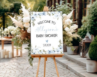 May include: A welcome sign for a baby shower with blue flowers and greenery. The sign says "Welcome to Allyson's Baby Shower" with a blue heart and the date 5.18.2024.