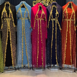 Könnte beinhalten: Eine Reihe farbenfroher, paillettenbesetzter Kleider auf Schaufensterpuppen. Die Kleider sind in Schwarz, Blau, Pink und Rot gehalten und mit goldenen Verzierungen versehen. Die Kleider haben lange Ärmel und sind bodenlang. Im Hintergrund steht der Text "SAYIN KAFTA".