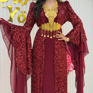 Könnte beinhalten: Ein bodenlanges bordeauxrotes Kleid mit Paillettenbesatz, langen, ausgestellten Ärmeln und einem Saum aus transparentem Chiffon. Das Kleid wird durch eine große, verzierte goldfarbene Halskette und einen passenden Metallgürtel mit Münzanhängern ergänzt.