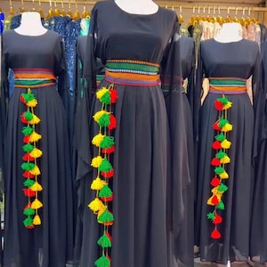 Può includere: Abito nero con maniche lunghe e cinturini ricamati colorati. Ogni abito presenta una lunga nappa di filo rosso, giallo e verde. Gli abiti sono esposti su manichini.