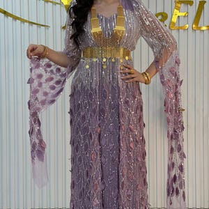 Könnte beinhalten: Ein bodenlanges lavendelfarbenes Kleid mit langen, fließenden Ärmeln, die mit blattförmigen Applikationen und silbernen Pailletten verziert sind. Das Kleid wird durch einen breiten, goldfarbenen Taillengürtel und eine passende Goldhalskette ergänzt.