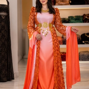 Könnte beinhalten: Ein formelles Kleid in Rosa- und Orangetönen. Das Kleid hat einen langen, fließenden Rock und eine passende Jacke mit aufwendigen, durchbrochenen Details. Ein goldener Gürtel mit Münzakzenten betont die Taille. Das Outfit wird durch einen passenden Schal ergänzt.