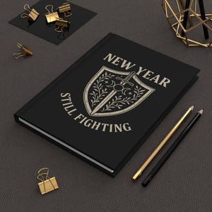 Può includere: Un diario nero con le parole "NEW YEAR STILL FIGHTING" e un disegno a scudo con una spada, mezzelune e stelle. Sono visibili anche matite dorate e nere e fermagli dorati.