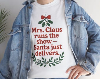 Könnte beinhalten: Weißes T-Shirt mit dem Text "Mrs. Claus runs the show - Santa just delivers." in Rot, Grün und Weiß. Das Design umfasst eine rote Schleife, Mistelzweige und einen grünen Kranz. Das Shirt wird mit einer blauen Jeansjacke getragen.