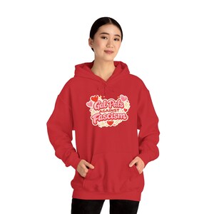May include: A red hoodie featuring the slogan "Gal Pals Against Fascism" in a heart-shaped design. The graphic includes pink and red hearts and a white border. The sweatshirt has a front pocket and a drawstring hood.