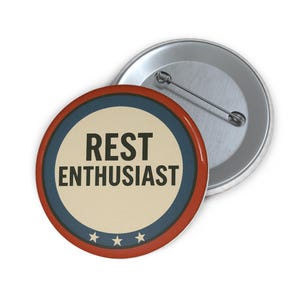 May include: A round button with the text "REST ENTHUSIAST" in black, set against a cream background, encircled by a blue ring, a red border, and three small stars. A second, unattached button is visible.