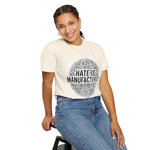May include: Cream-colored t-shirt with the text "HATE IS MANUFACTURED" inside a black and white gear design. The shirt is short-sleeved and worn by a person wearing blue jeans.