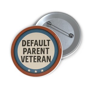 以下が含まれることがあります： 黒色の「DEFAULT PARENT VETERAN」の文字が入った丸いボタン。ボタンはクリーム色の中心、青いリング、茶色の外側のリングで構成されています。下部には3つの白い星があります。2つ目の銀色のボタンが後ろにあります。