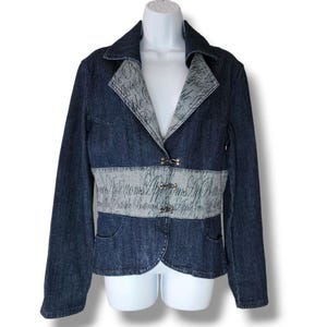 May include: A dark wash denim jacket with a light grey accent on the lapel and waist. The light grey accents feature the word "Apple Bottoms" in a script font. The jacket has two decorative clasps and two front pockets.