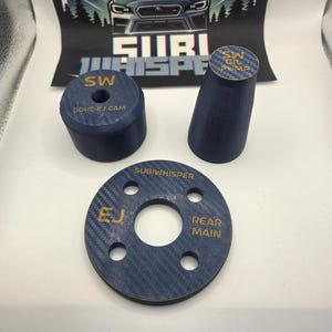 Op de afbeelding: Een assortiment donkerblauwe auto-onderdelen met gouden tekst, waaronder een cirkelvormig onderdeel met "SUBIWHISPER", "EJ" en "REAR MAIN" erop. Andere onderdelen zijn een cilinder en een kegelvormig onderdeel. Een Subaru-logo is op de achtergrond.