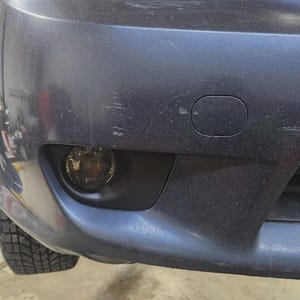 May include: Close-up of a dark blue car bumper with a fog light and a tire visible. The bumper has scratches and a small, oval-shaped cover. The fog light is round and has a yellow tint.