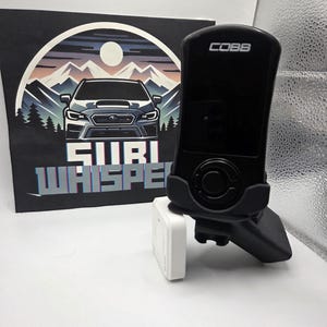 May include: A black Cobb tuning device with a black mounting bracket and a white rectangular device. Behind the devices is a black sign with a car graphic, mountains, and the text "SUBI WHISPER".