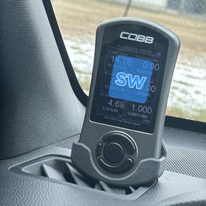 May include: A black COBB tuning device displaying real-time engine data, mounted in a car's dashboard. The screen shows numbers and text, including "14.78" and "4.69". The device is held in a gray plastic holder.