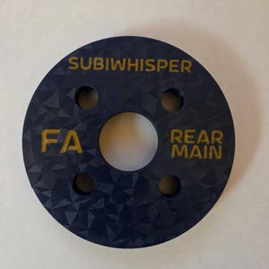 Op de afbeelding: Een cirkelvormig donkerblauw onderdeel met een geometrische, gefacetteerde oppervlaktestructuur. De tekst SUBIWHISPER staat bovenaan, met FA links en REAR MAIN rechts in het geel. Het heeft een groot middengat en vier kleinere gaten.