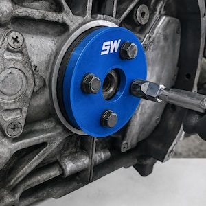Op de afbeelding: Een close-up van een motoronderdeel met een helderblauwe, cirkelvormige component. De blauwe component heeft de letters "SW" in wit en is vastgezet met bouten. Een gereedschap wordt gebruikt om aan de component te werken.