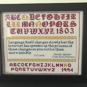 May include: A framed cross-stitch piece with a white background and red, yellow, and blue lettering. The piece features the alphabet in a traditional font, the years 1803 and 1994, and a quote about language change. The quote reads: "Language itself changes slowly but the internet has speeded up the process of those changes so you notice them more quickly."  D. Crystal. Spanish alphabet From 29 to 27 letters.