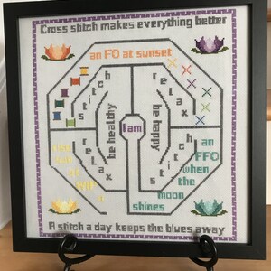 May include: A framed cross-stitch design with a white background and purple and gray stitching. The design features a circular pattern with the words "I am healthy, I am happy, an FO at sunset, a stitch a day keeps the blues away" and other words and symbols.