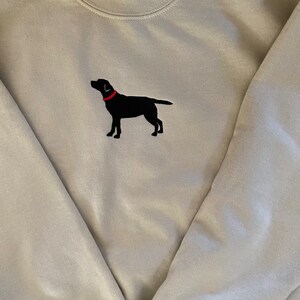 May include: A light beige sweatshirt with a black embroidered Labrador Retriever wearing a red collar.