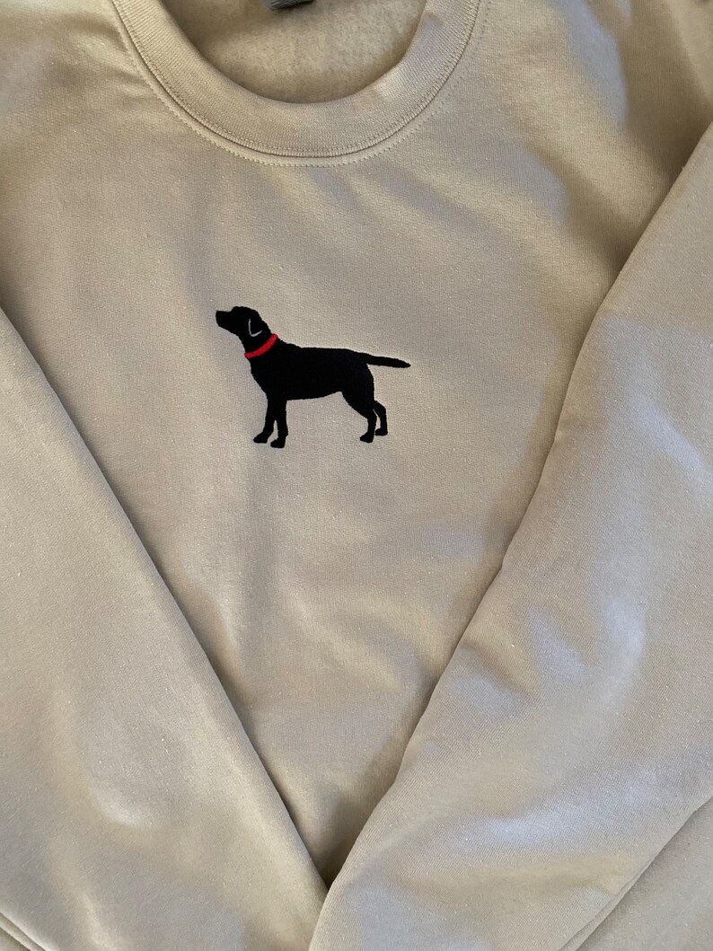 May include: A light beige sweatshirt with a black embroidered Labrador Retriever wearing a red collar.