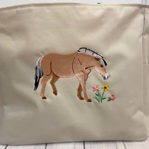 Può includere: Una borsa tote in tela beige con un'immagine ricamata di un cavallo marrone con criniera e coda bianche, che pascola sull'erba verde con fiori rosa e gialli.