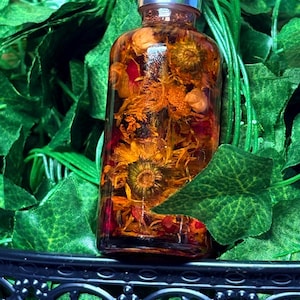 May include: A clear glass bottle filled with dried flowers and herbs, topped with a silver dropper. The bottle is surrounded by green leaves and sits on a black, ornate surface. The contents of the bottle appear to be a golden-orange color.