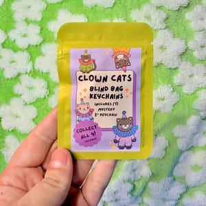 Peut inclure: Un sachet aveugle jaune pour porte-clés avec le texte "CLOWN CATS" et des illustrations de chats en costumes de clown. Le paquet comprend un porte-clés mystère de 5 cm. Le texte "COLLECT ALL 4!" est également visible.