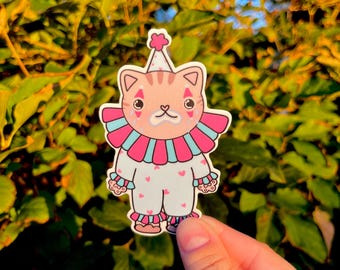 May include: A sticker featuring a cartoon cat dressed as a clown. The cat has a pink and blue ruffled collar, a white jumpsuit with pink hearts, and a party hat. The cat's face has a sad expression with teardrops.