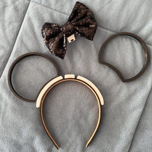 Puede incluir: Un lazo marrón con lentejuelas y dos recortes marrones con forma de oreja se muestran con una diadema con un diseño blanco y beige. Los artículos están dispuestos sobre una superficie azul claro texturizada, lo que sugiere un tema de accesorios.