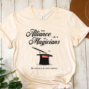 May include: A beige t-shirt with a black graphic of a top hat and magic wand. The text "The Alliance of Magicians" is written in a stylized font above the graphic. The text "We demand to be taken seriously" is written below the graphic.