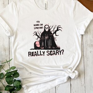 May include: White t-shirt with a black and white graphic of a spooky figure with a skull face and the text "You wanna see something really scary?"