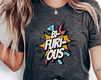 May include: Dark gray t-shirt with the text "BI-FURIOUS" in a comic book style speech bubble. The bubble has yellow, red, and blue accents. The shirt is a crew neck style.