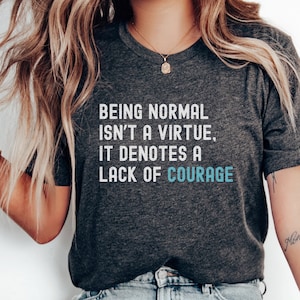 May include: A gray t-shirt with the text "Being normal isn't a virtue, it denotes a lack of courage" printed in white. The text "More Ocean" is tattooed on the wearer's arm.