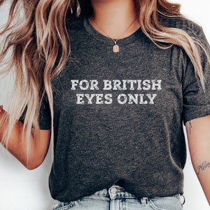 May include: Dark gray V-neck t-shirt with the text "FOR BRITISH EYES ONLY" in white, centered on the chest. The shirt is worn by a person with long, wavy blonde hair and a tattoo on the arm.