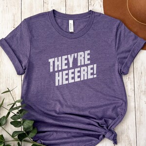 K&ouml;nnte beinhalten: Lilafarbenes T-Shirt mit kurzen &Auml;rmeln und dem Text "They're Heeeere!" in wei&szlig;en Buchstaben.