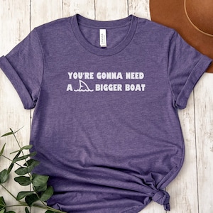 May include: A purple t-shirt with the text "You're gonna need a bigger boat" and a shark fin graphic printed in white.