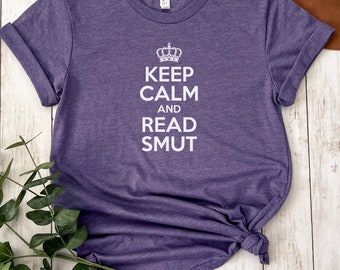 May include: Purple t-shirt with rolled sleeves and the text "KEEP CALM AND READ SMUT" in white, topped with a crown graphic. The shirt is knotted at the bottom. A brown hat and greenery are in the background.