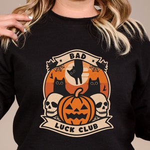 May include: Black sweatshirt featuring a Halloween-themed graphic. The design includes a pumpkin, black cats, skulls, and the text "BAD LUCK CLUB" in a banner. The background has an orange and tan color scheme.
