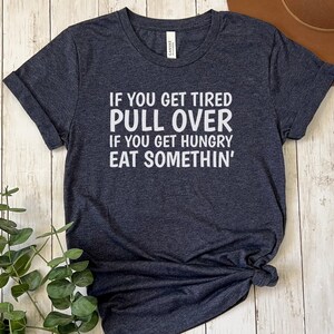May include: A dark heather grey t-shirt with white text that reads "If you get tired pull over if you get hungry eat somethin'"