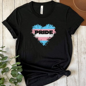 May include: Black t-shirt with a distressed heart design in the colors of the transgender pride flag. The heart has the word "PRIDE" written across it.