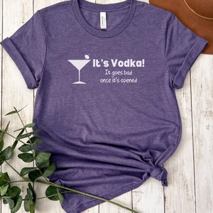May include: Purple short-sleeved t-shirt with a white martini glass graphic and the text "It's Vodka! It goes bad once it's opened."