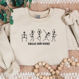 May include: A beige sweatshirt with a graphic of five skeletons dancing and the text "Rattle Dem Bones".