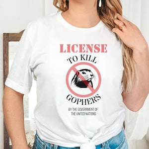 May include: White t-shirt with the text "LICENSE TO KILL GOPHERS" in pink and black. The design includes a gopher image inside a red circle with a slash, and the text "BY THE GOVERNMENT OF THE UNITED NATIONS".