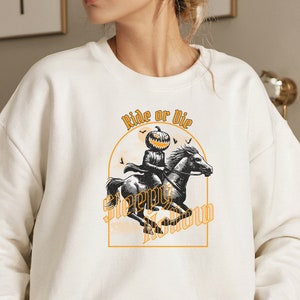 May include: Cream-colored sweatshirt featuring a Halloween-themed graphic. The design includes a pumpkin-headed figure riding a horse, with the words "Ride or Die" and "Sleepy Hollow" in an orange arch. Black bats are also included in the design.