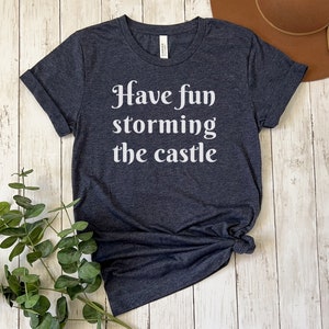 May include: A dark heather grey t-shirt with the text "Have fun storming the castle" printed in white.