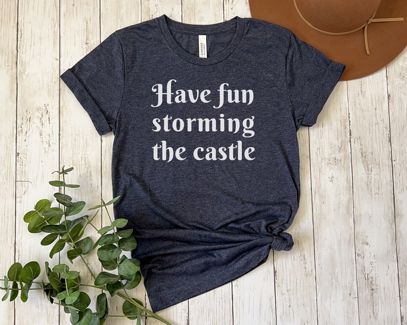May include: A dark heather grey t-shirt with the text "Have fun storming the castle" printed in white.