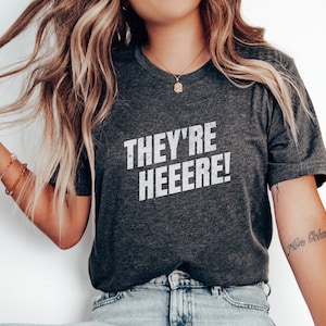 K&ouml;nnte beinhalten: Ein dunkelgraues T-Shirt mit dem Aufdruck "THEY'RE HEEEERE!" in Wei&szlig;. Das Shirt wird von einer Person mit langen blonden Haaren getragen.