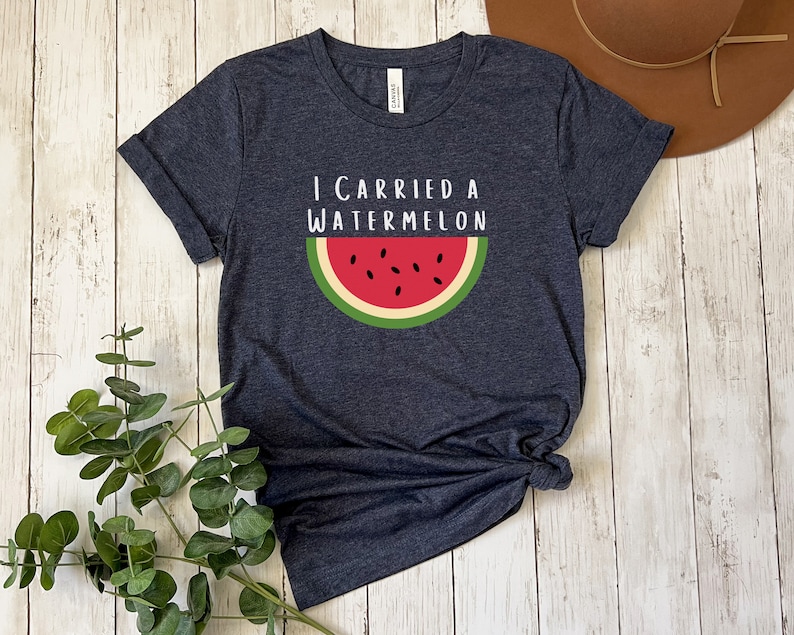 Może przedstawiać: Koszulka w ciemnoniebieskim kolorze z napisem "I carried a watermelon" i grafiką plasterka arbuza z czerwonym miąższem i czarnymi pestkami.