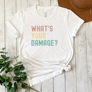 May include: White t-shirt with the text "WHAT'S YOUR DAMAGE?" in a retro color scheme. The text is in a stacked format with each word in a different color.